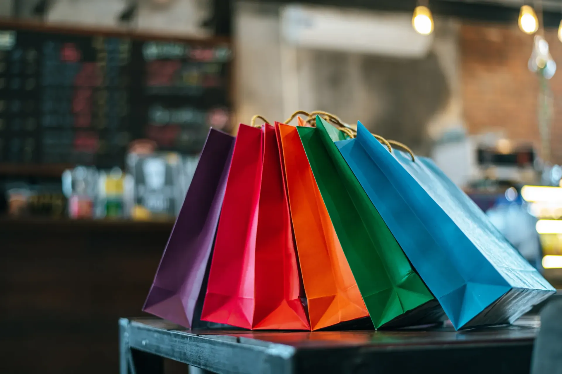 A row of colorful shopping bags in shades of purple, red, orange, green, and blue standing on a table.