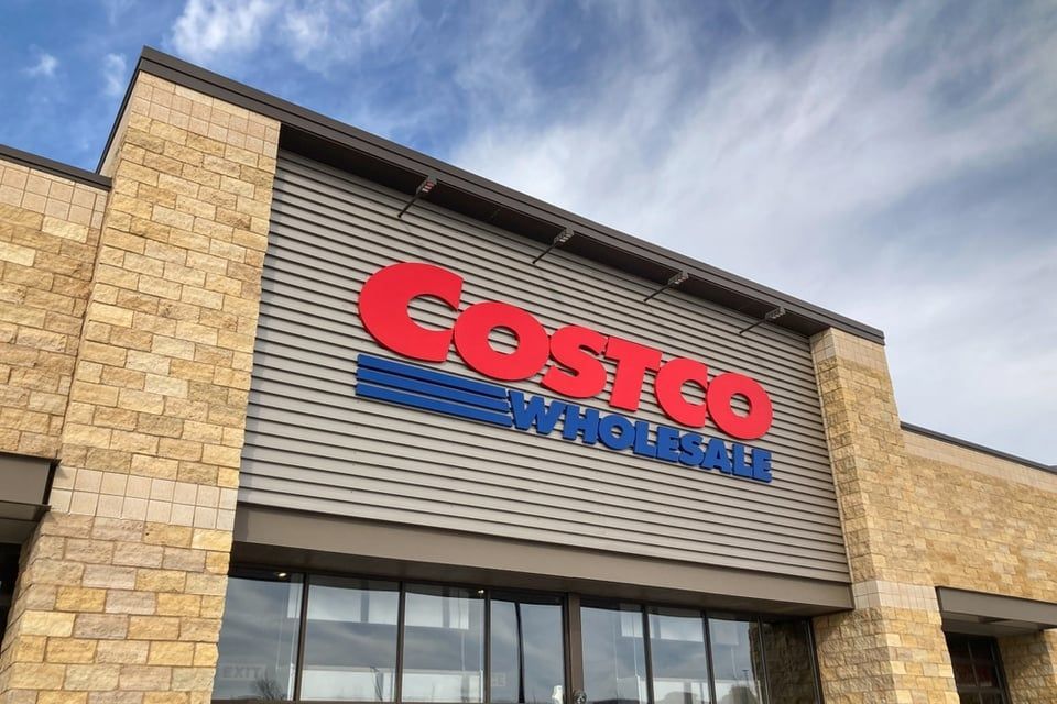 The exterior of a Costco Wholesale store featuring a red and blue logo on a grey, corrugated metal facade and stone walls.
