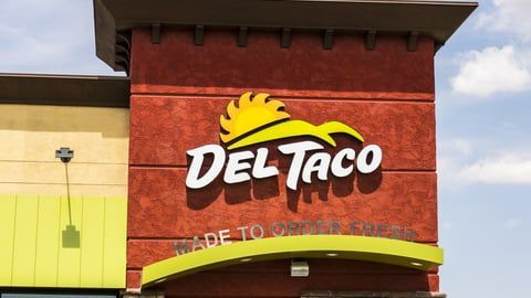 The red brick storefront of a Del Taco restaurant with a yellow logo, set against a blue sky.