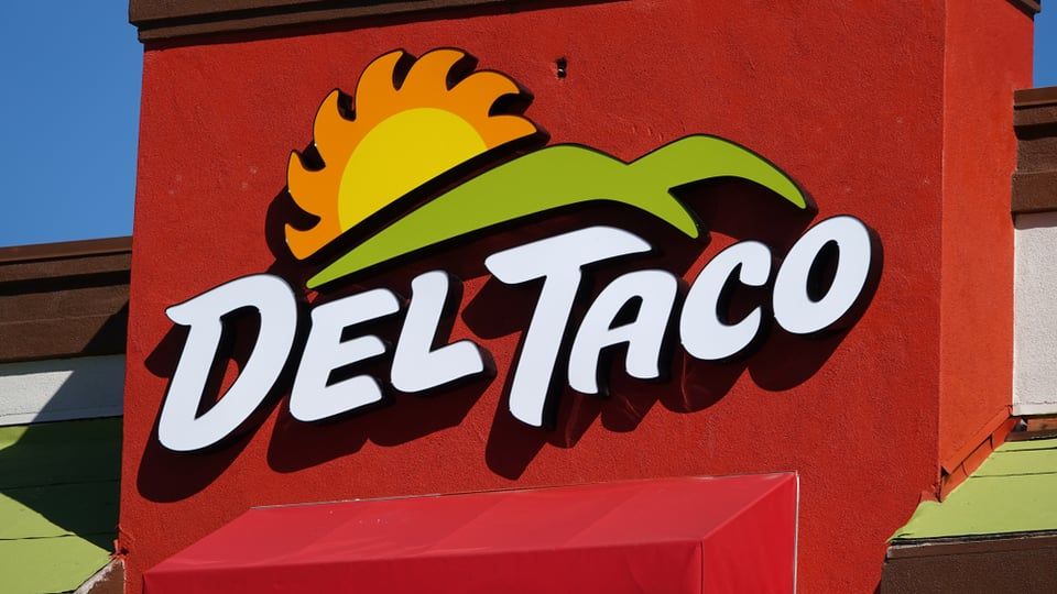 The white Del Taco logo featuring a stylized yellow sun and green hill is mounted on a vibrant red wall.