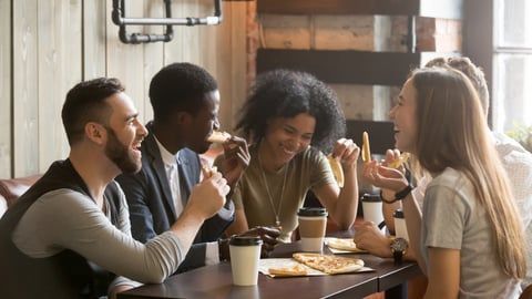 Four friends laughing and eating pizza together at a cafe table with coffee cups.