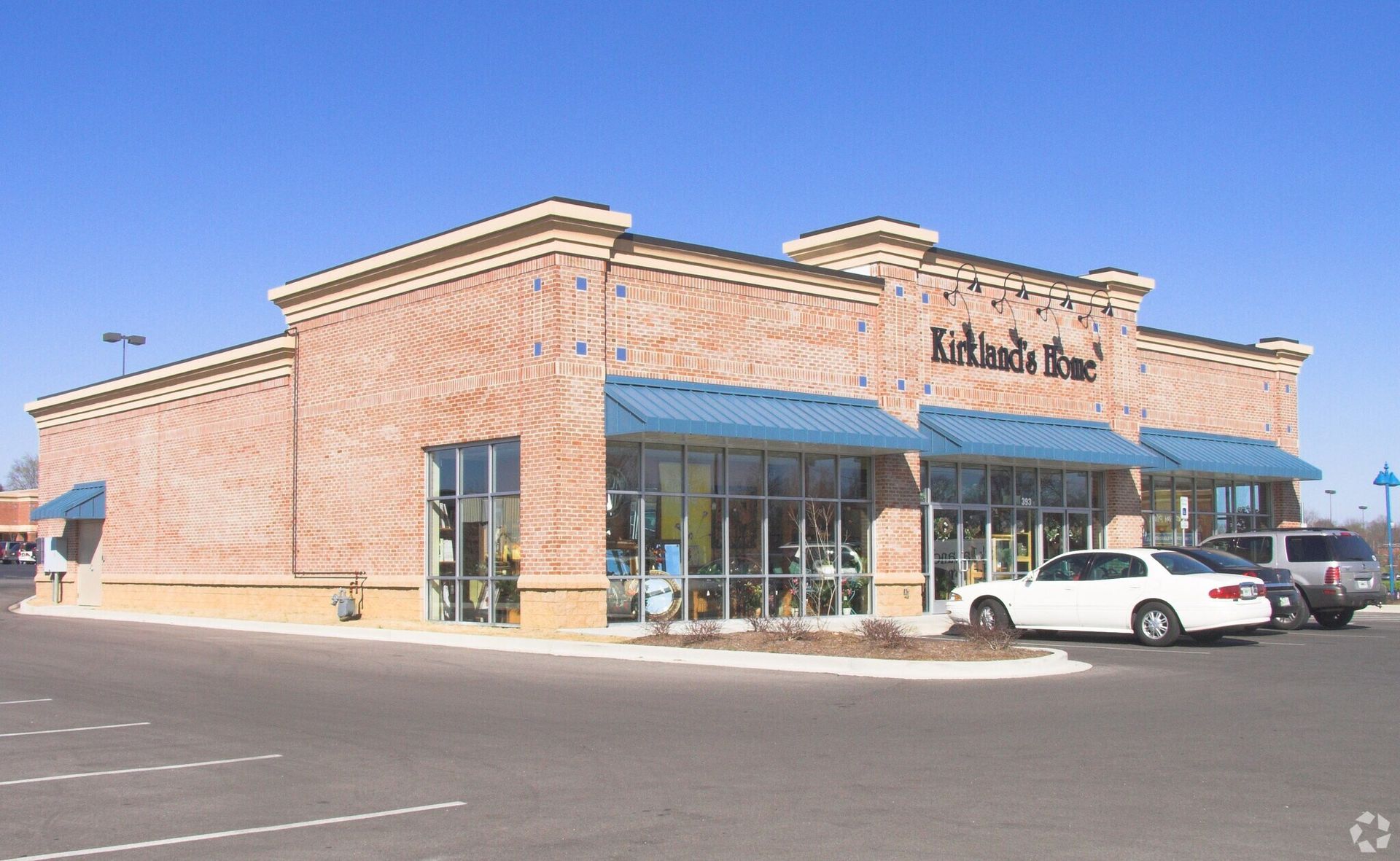 A Kirkland's Home store in a brick-faced retail strip center under a clear blue sky, with a parking lot in the foreground.