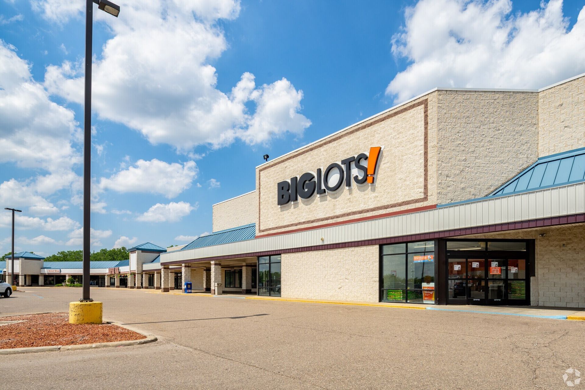 A tan, stone-faced Big Lots store exterior in a shopping center under a blue sky with fluffy white clouds.