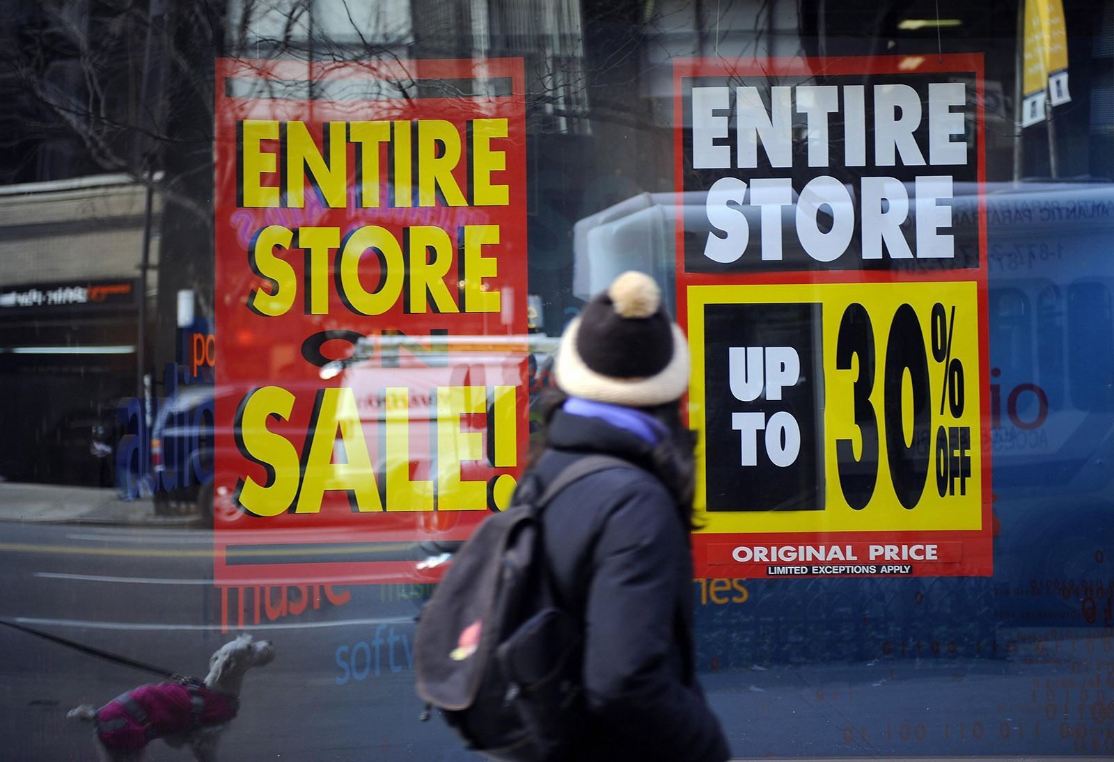 A person with a backpack walks past a shop window with red and yellow signs advertising an 