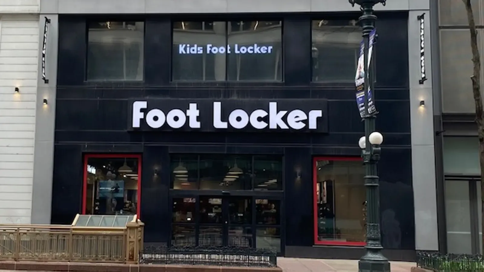 The storefront of a Foot Locker and Kids Foot Locker with black walls, red accents, and large windows on a city street.