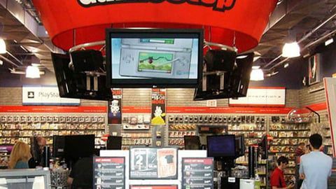 Interior view of a GameStop store with game displays on shelves, a central monitor, and employees at the service desk.