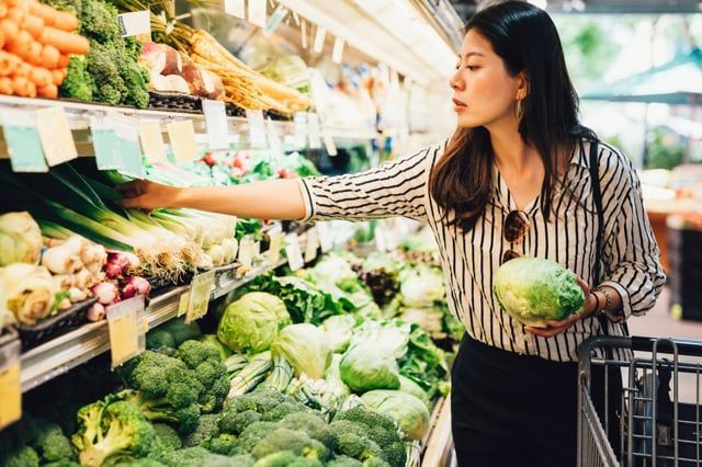 A person shops for fresh produce in a grocery store, holding a head of cabbage and reaching for green vegetables on a shelf.