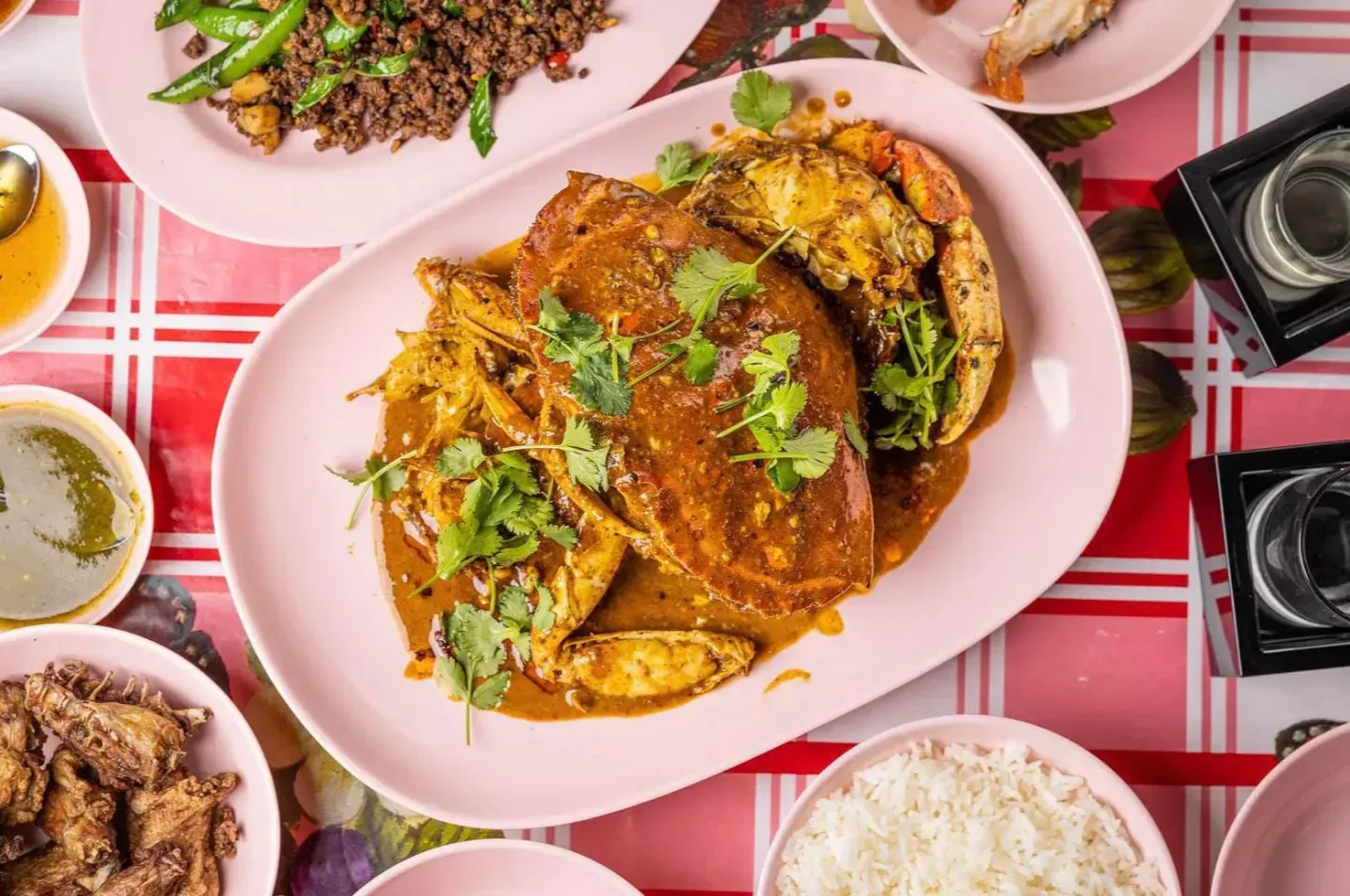 A high-angle shot of various Thai dishes served on pink plates atop a red and white checkered tablecloth.