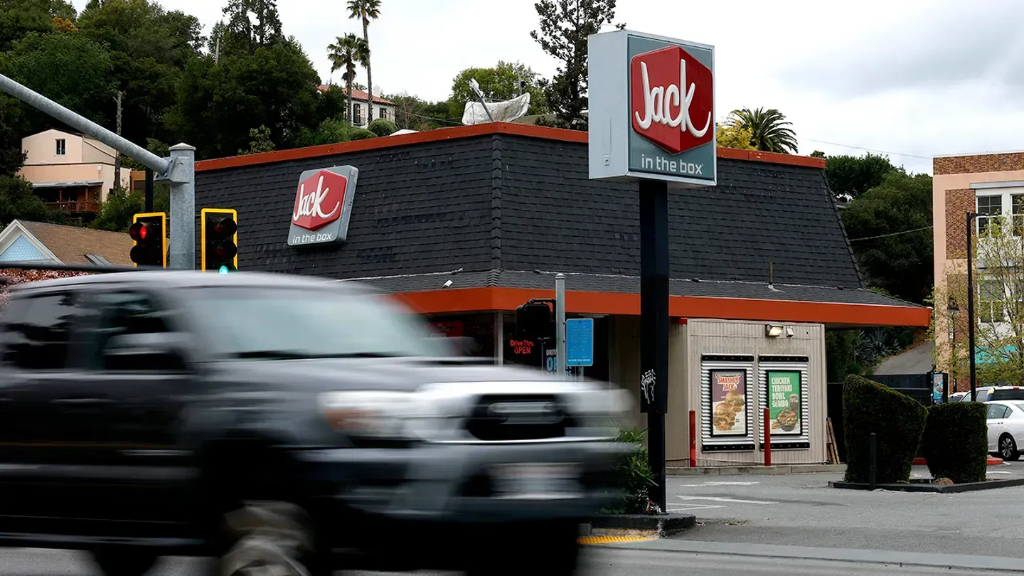 A motion-blurred black truck drives past a Jack in the Box restaurant with a gray roof and sign on a cloudy day.