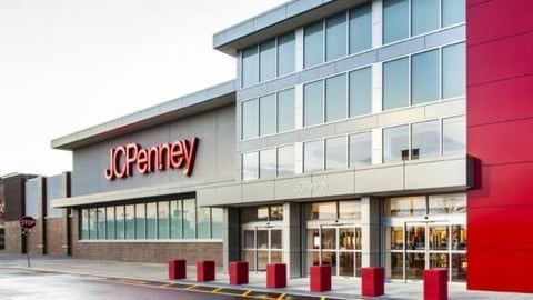 A modern JCPenney store exterior with a red accent wall, glass doors, and a line of red bollards in front.