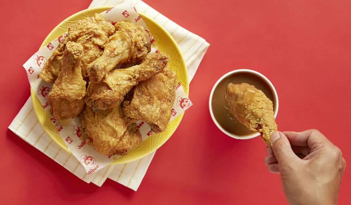 A hand dips a piece of golden-brown fried chicken into a small bowl of gravy next to a plate of chicken on a red surface.