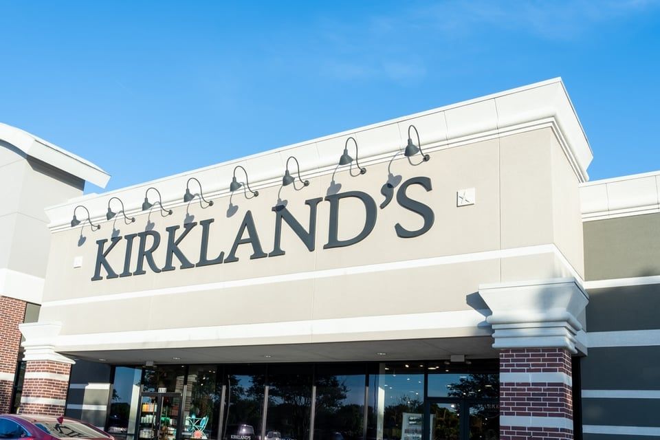 The exterior storefront of a Kirkland's home decor store with a beige facade, black lettering, and metal lighting fixtures.