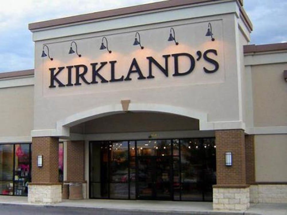 The entrance of a Kirkland’s home decor store with a beige facade, black lettering, and wall-mounted exterior lamps.