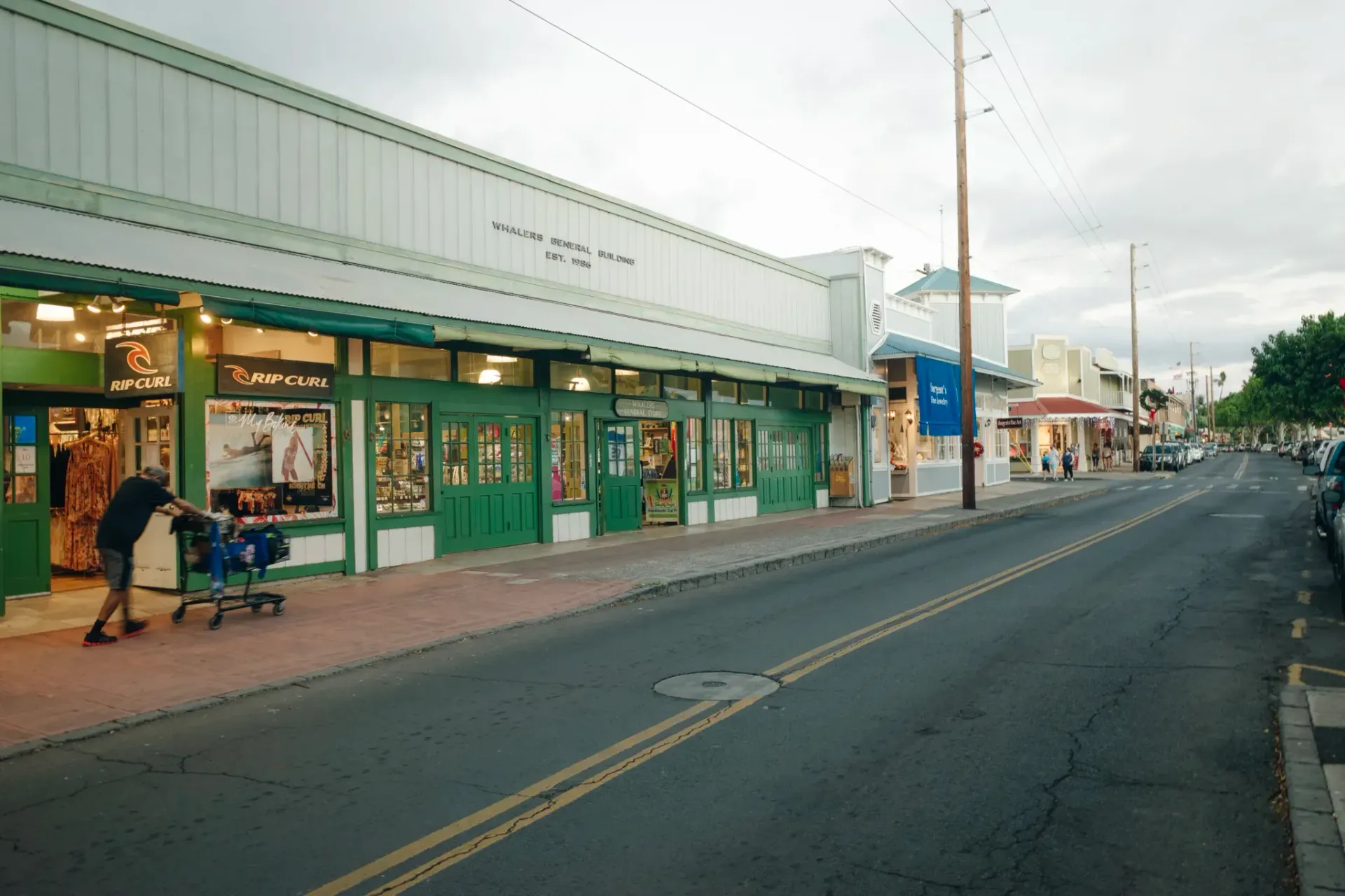 A person pushes a cart along the sidewalk of a street lined with storefronts featuring green trim and large windows.