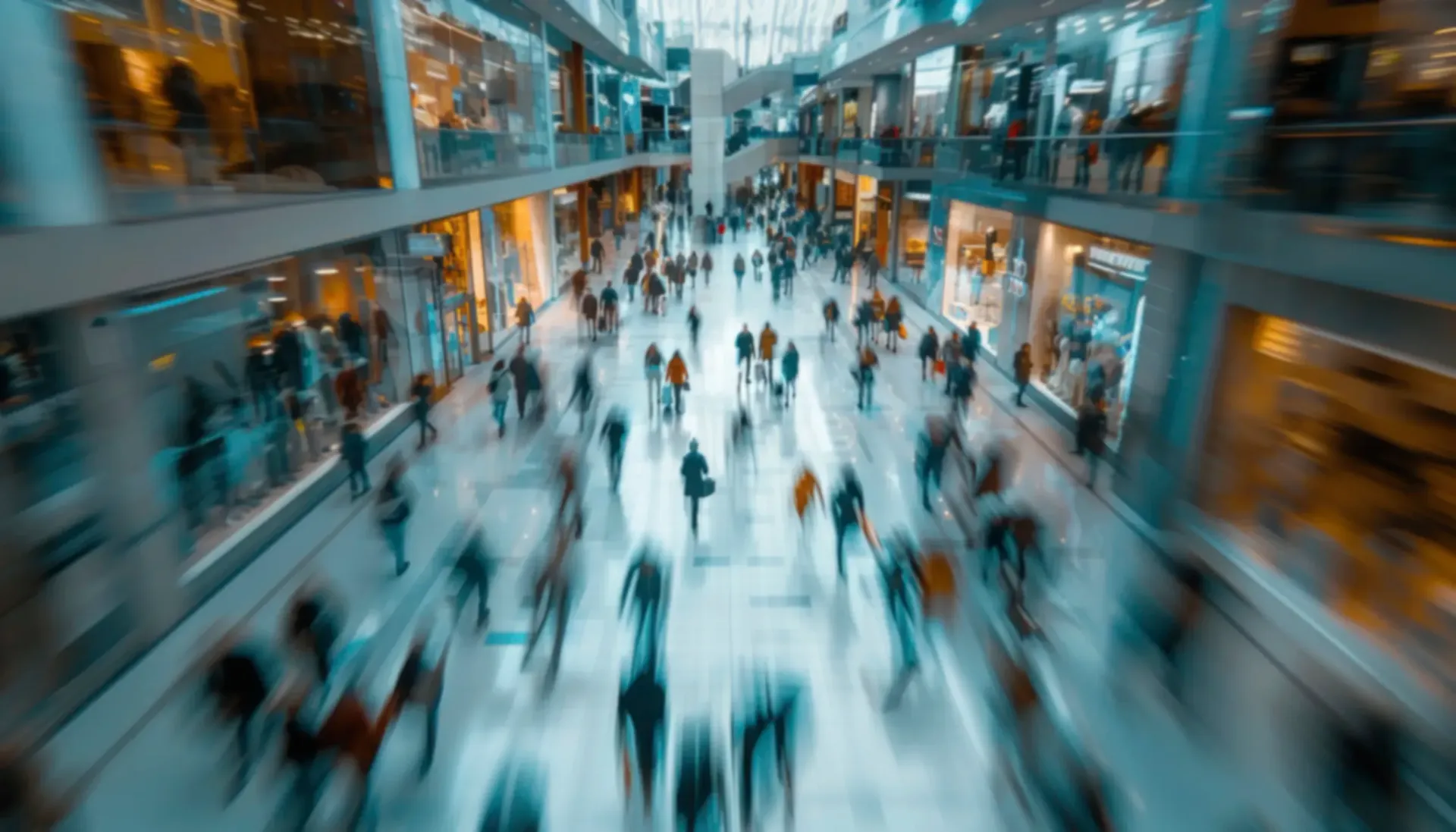 A motion-blurred, high-angle view of many people walking through a brightly lit, modern multi-level shopping mall.
