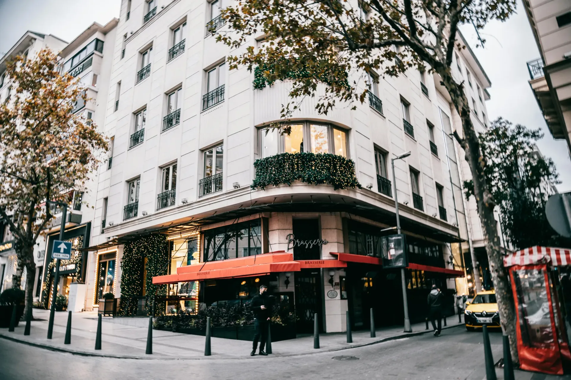 A street-level view of a white multi-story building featuring an outdoor cafe with red awnings on a tree-lined corner.