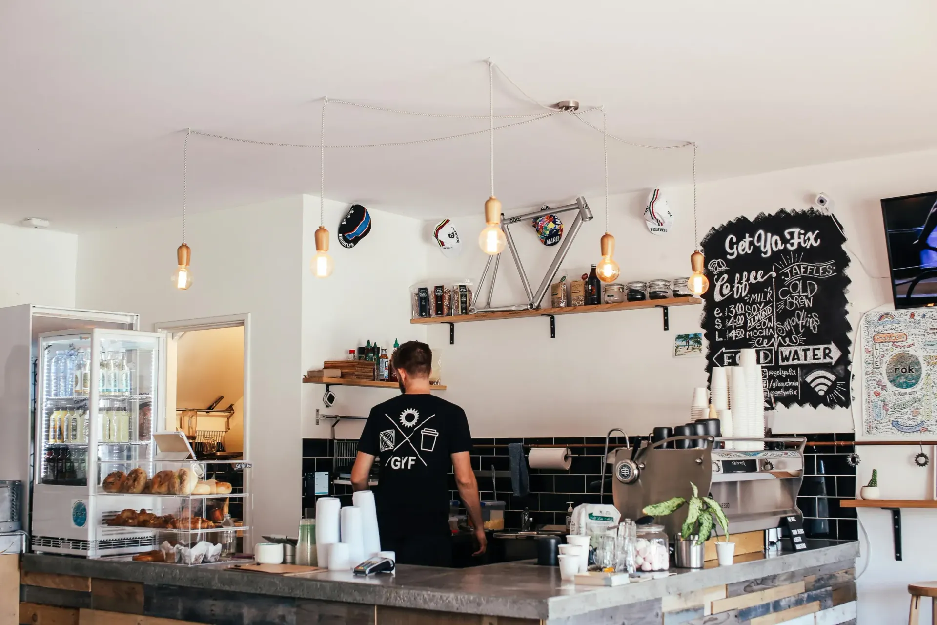 A person in a black t-shirt works behind the counter of a modern coffee shop with white walls and a chalkboard menu.
