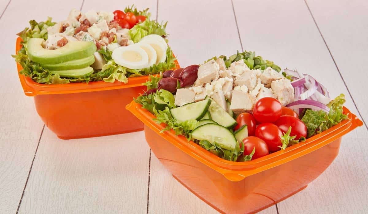 Two square orange containers filled with fresh salads topped with chicken, avocado, tomatoes, cucumber, and red onion.