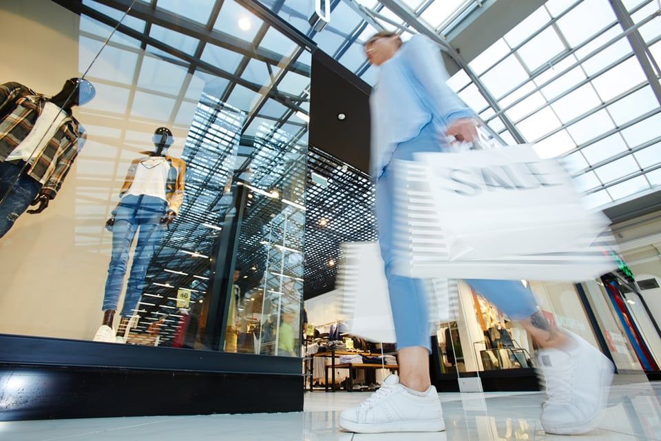 A shopper in blue clothes walks past a clothing store window display while holding several white shopping bags.