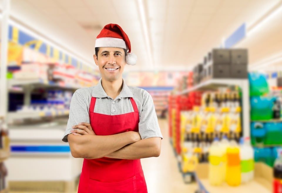 A smiling retail worker in a grey polo, red apron, and Santa hat stands with arms crossed inside a grocery store.