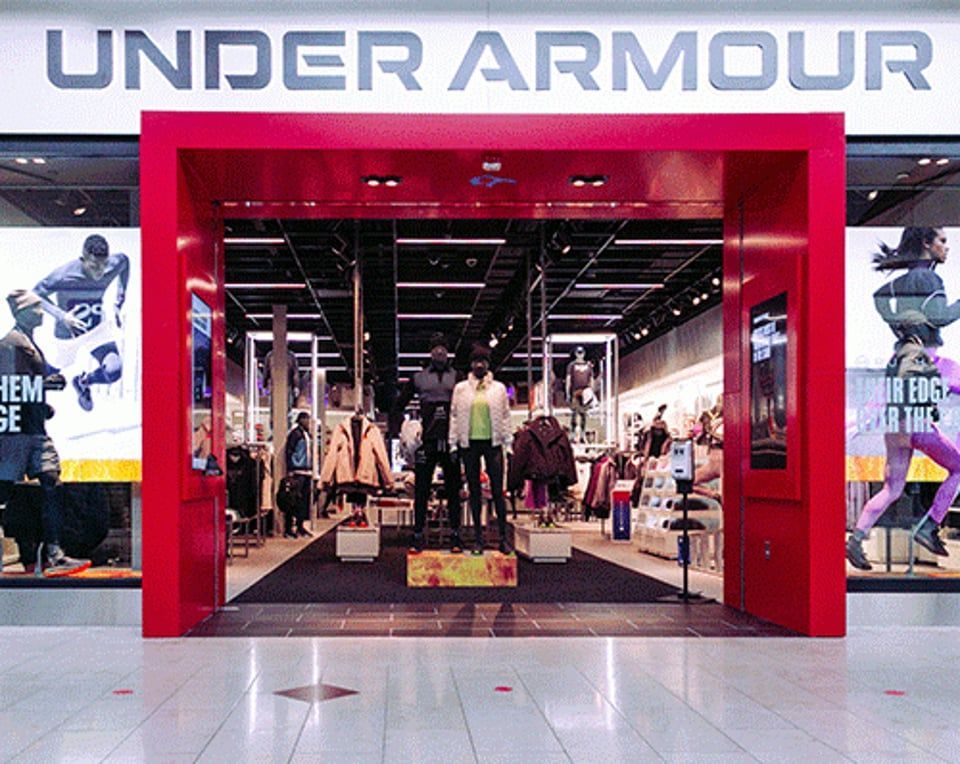 Storefront of an Under Armour shop with red entrance framing, large windows featuring athletic posters, and mannequins.