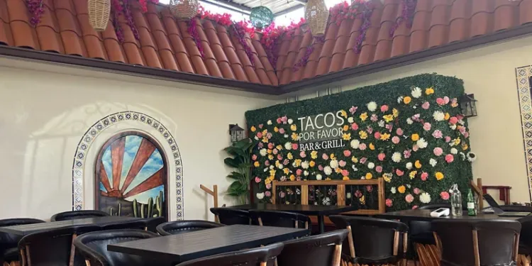 Interior view of a restaurant with a tiled roof, a sunrise wall mural, and a flower-covered wall with the sign Tacos.