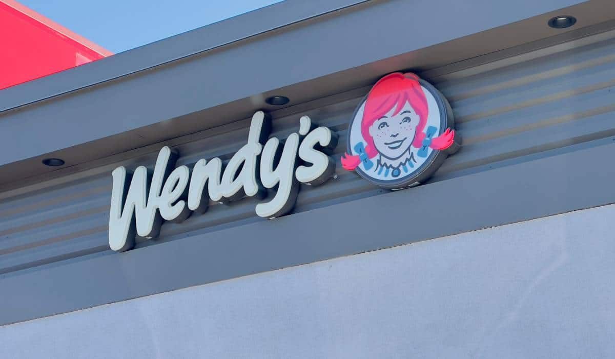 The Wendy's logo with the signature red-haired girl mascot mounted on a gray building exterior under a blue sky.