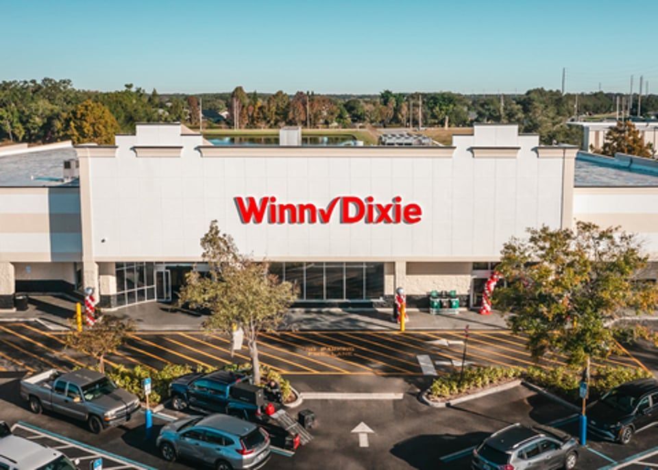 An aerial view of a white Winn-Dixie grocery store building with a parking lot in the foreground on a sunny day.