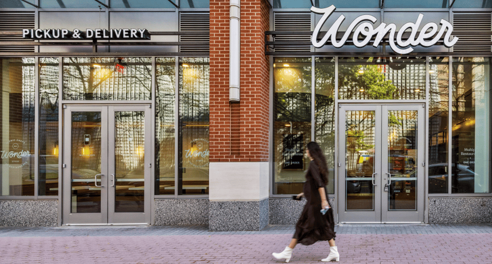 A woman in a dark dress and white boots walks on a sidewalk in front of a Wonder restaurant storefront.