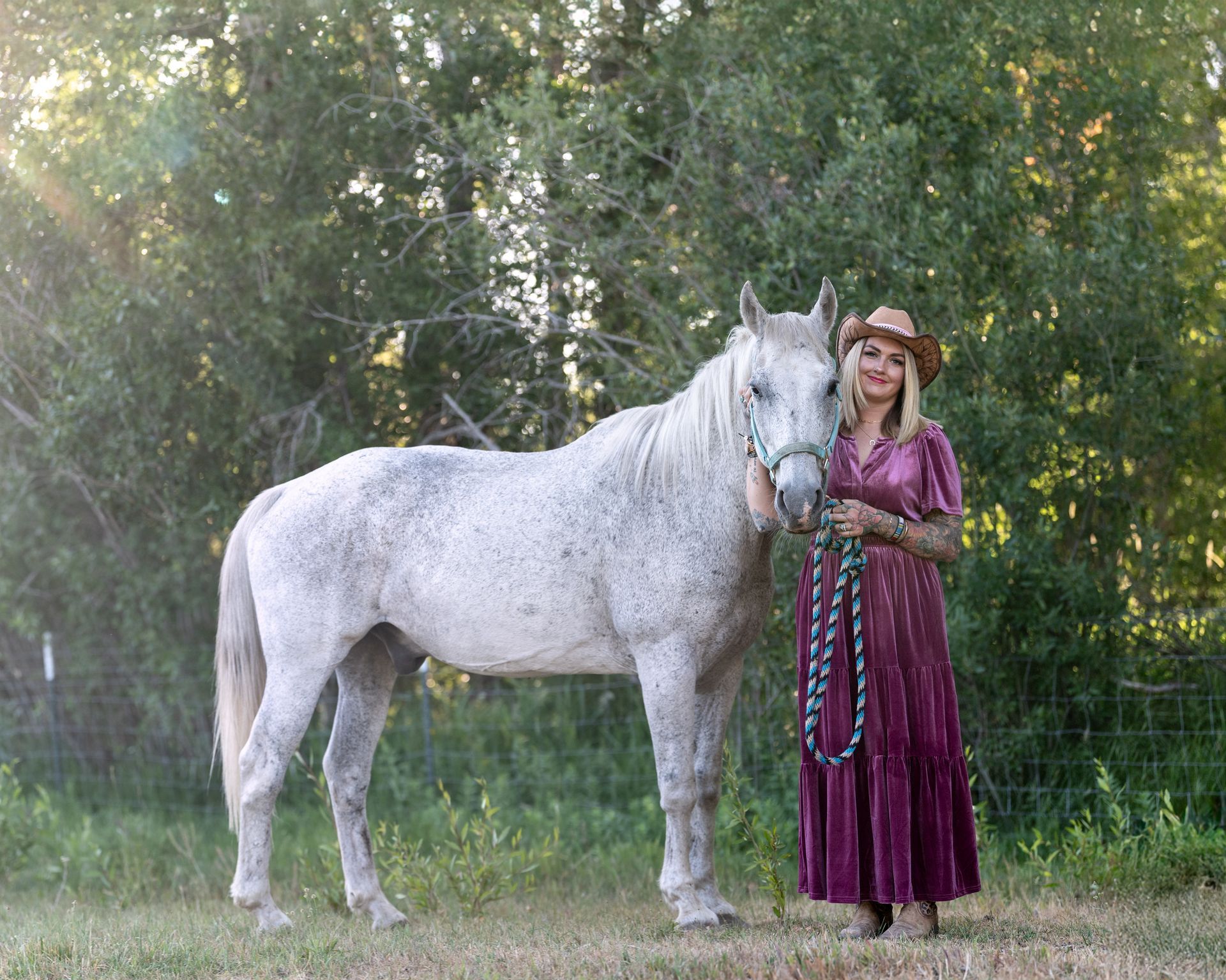Woman in a purple dress and hat standing with a white horse in a wooded field.