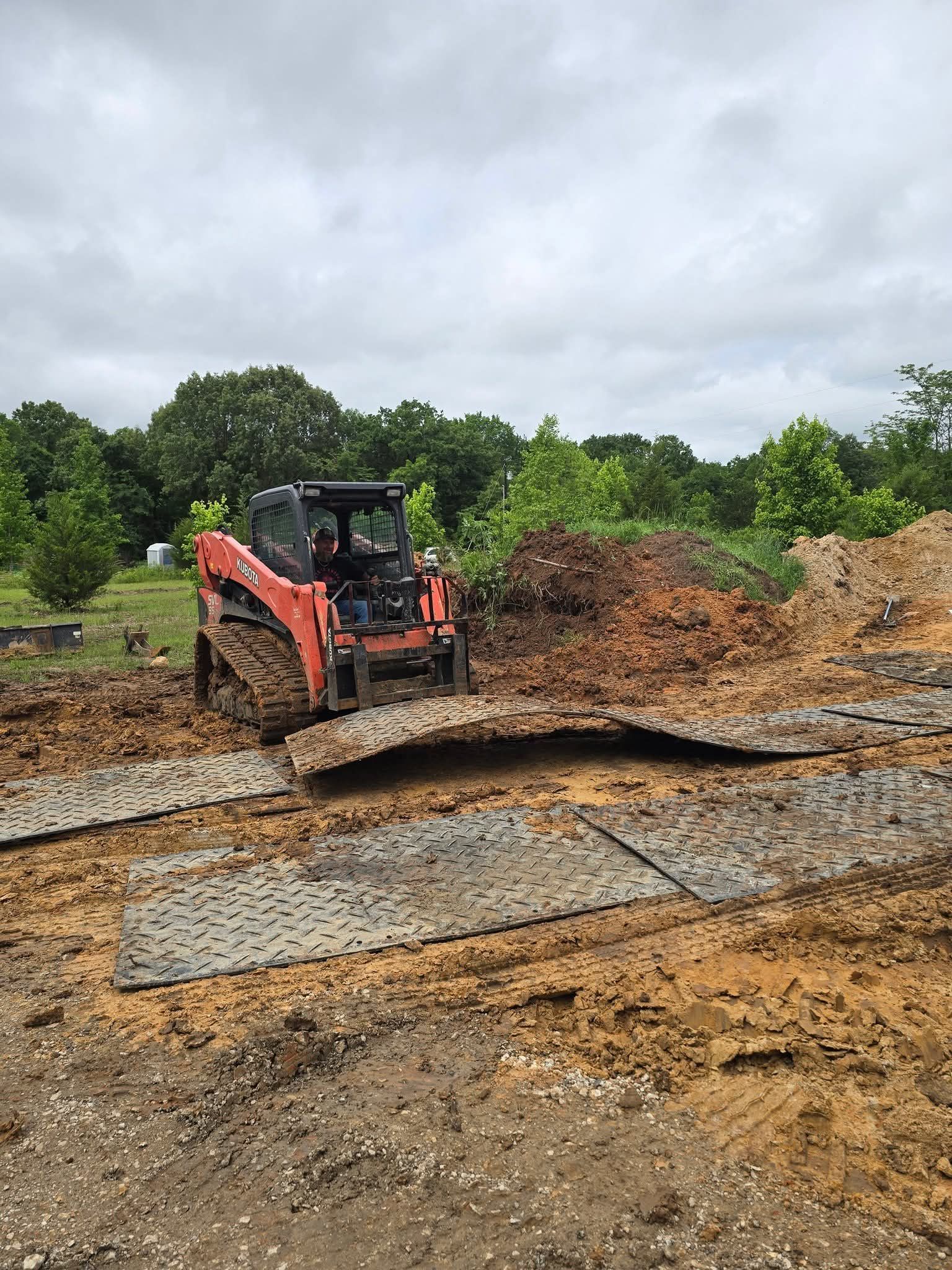 Orange skid steer on a muddy construction site, moving soil. Overcast sky and trees in the background.