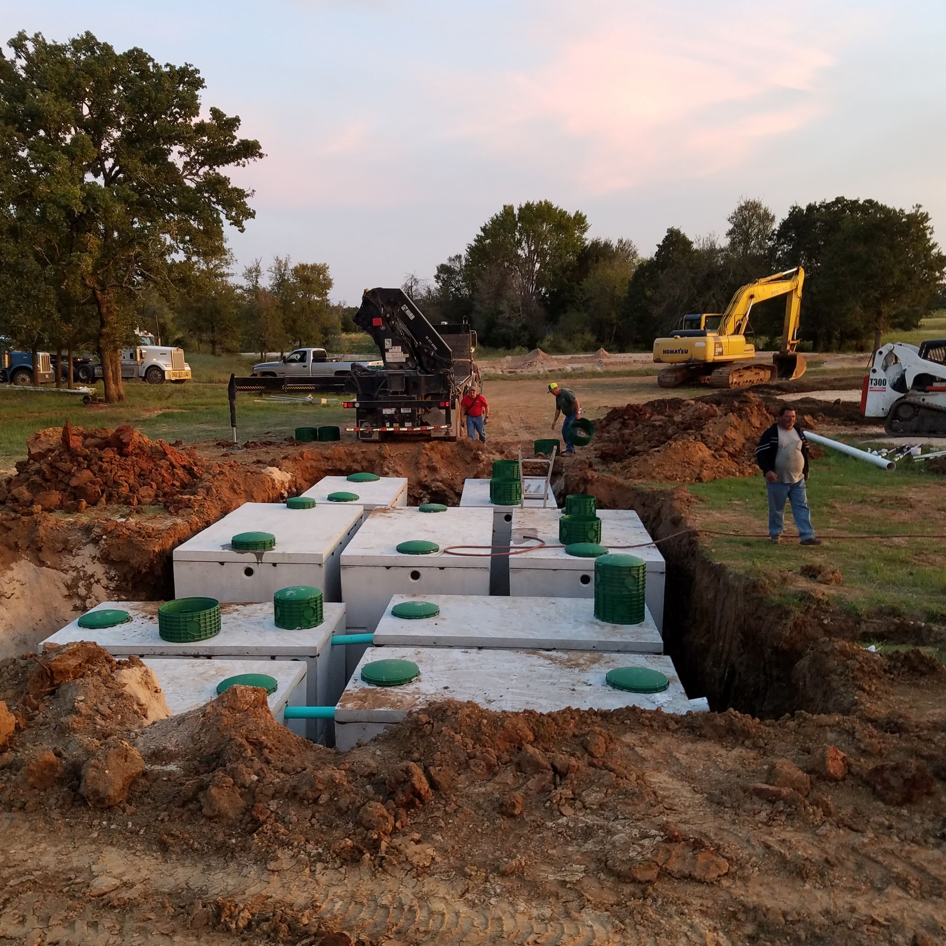 Construction site with septic tanks installed in a large excavated area; workers, crane, and excavator visible.