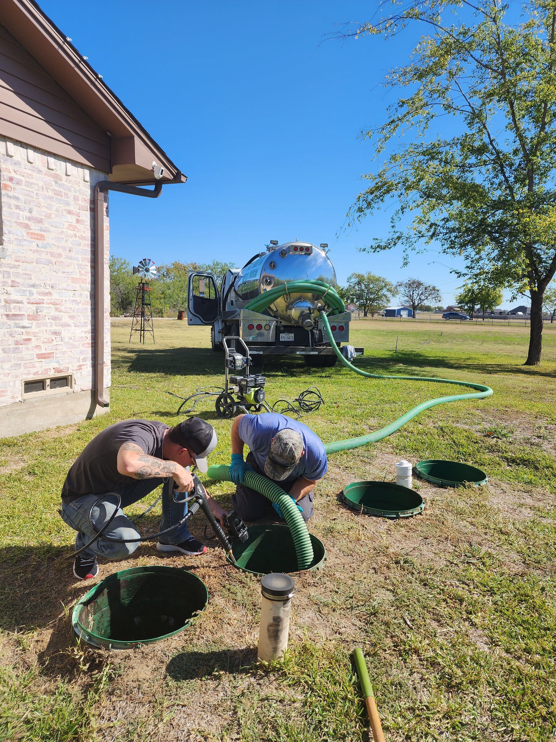 Two workers pumping a septic tank. A truck and hose are visible in a yard on a sunny day.