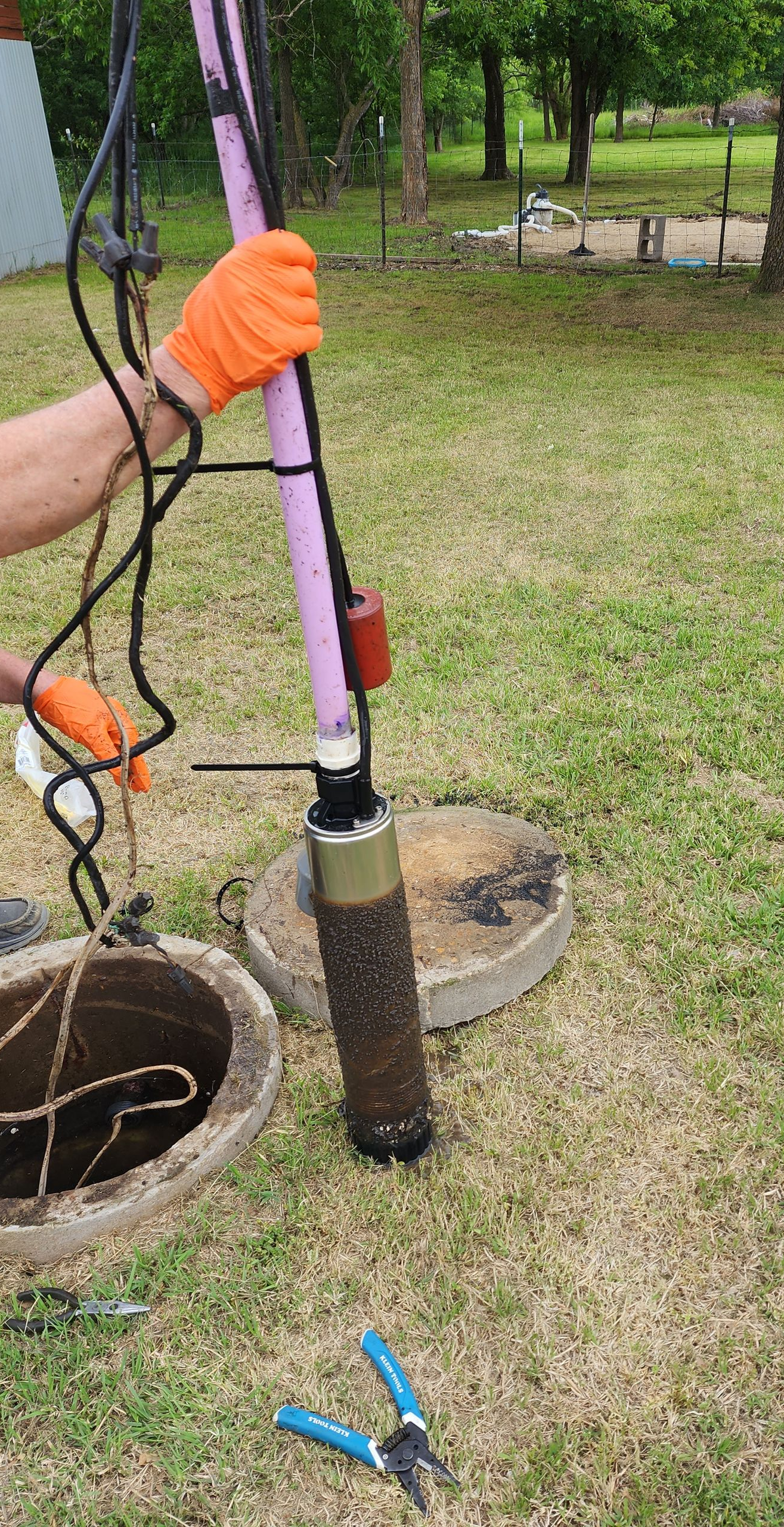 Person in orange gloves holding a well pump, removing it from a well in a grassy yard.