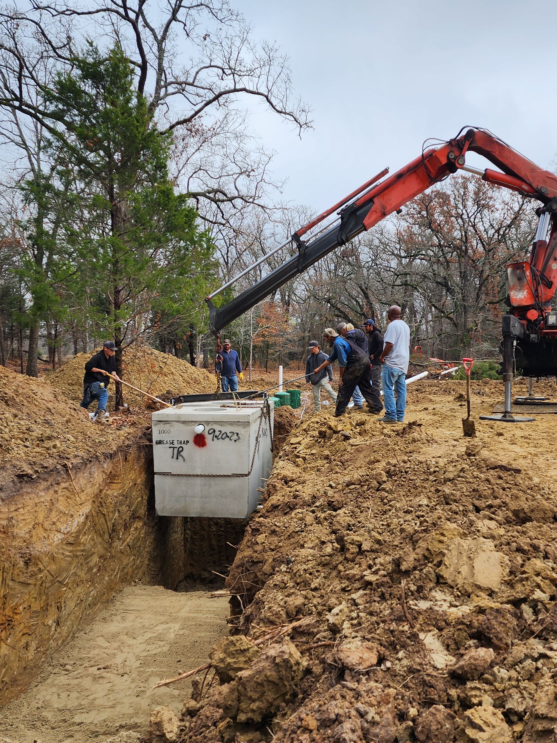 Crane lowering septic tank into trench; workers stand nearby. Earth-moving equipment present.