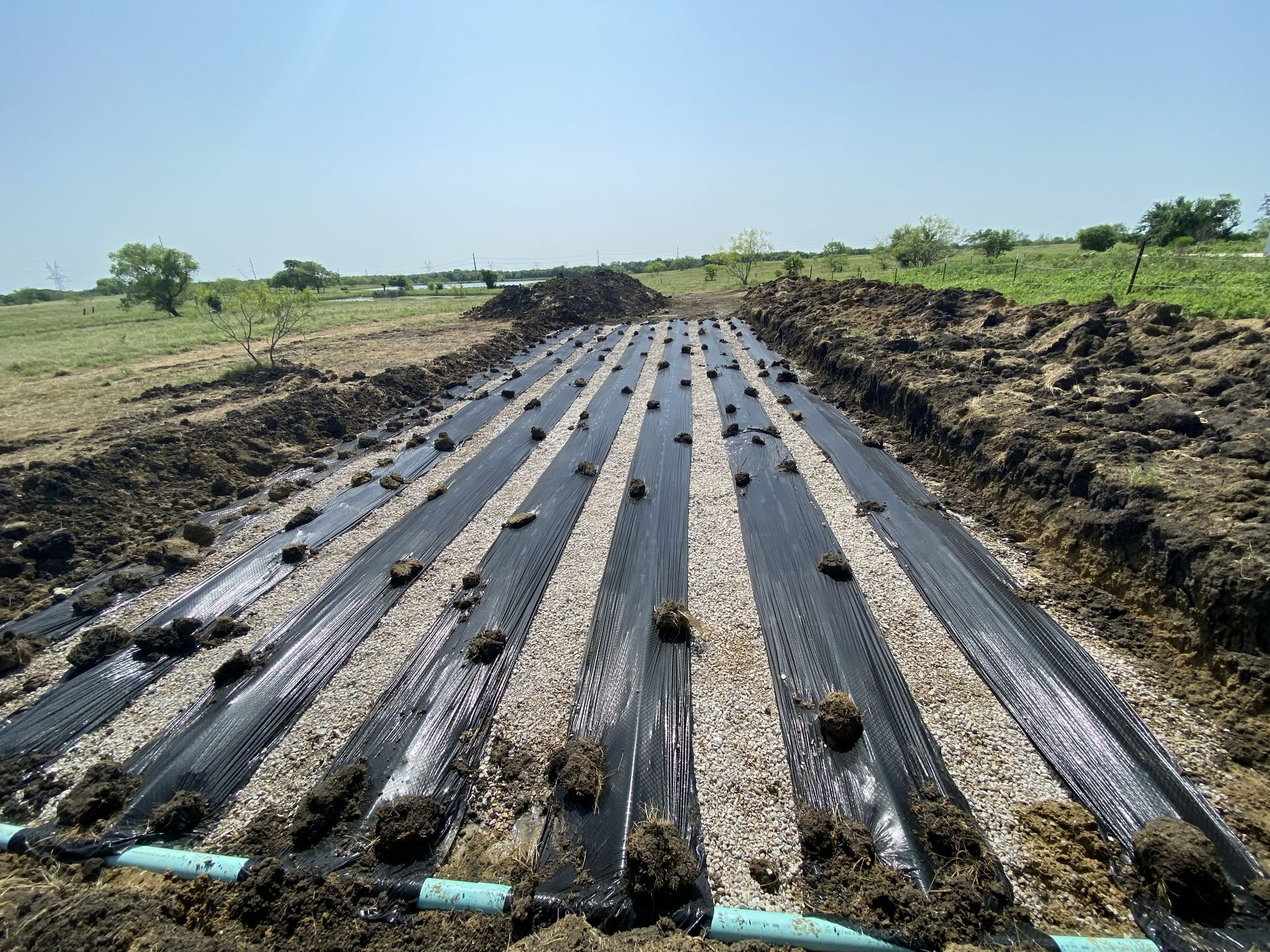 Black landscape fabric covers gravel-filled trenches with irrigation piping for a garden bed.