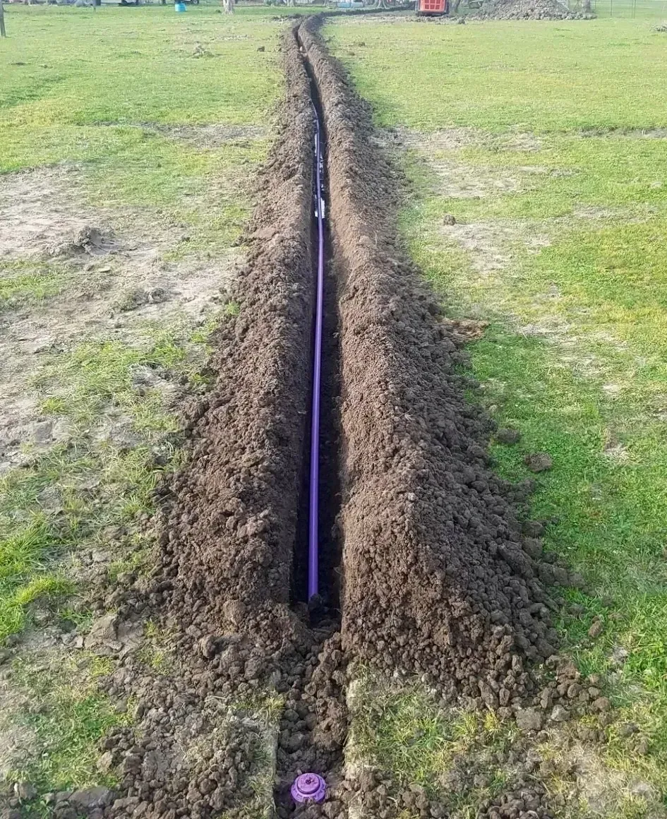 Trench dug in grass, holding a purple pipe, likely for utilities.