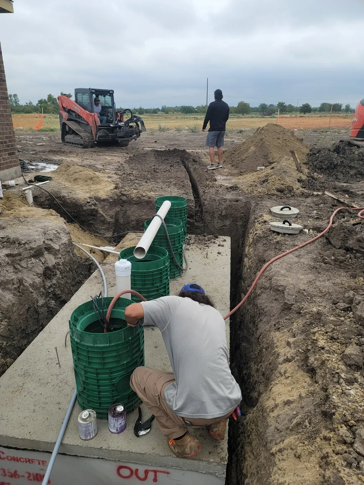 Construction worker inspecting septic system, in a trench. A skid steer and another worker are in the background. Cloudy day.