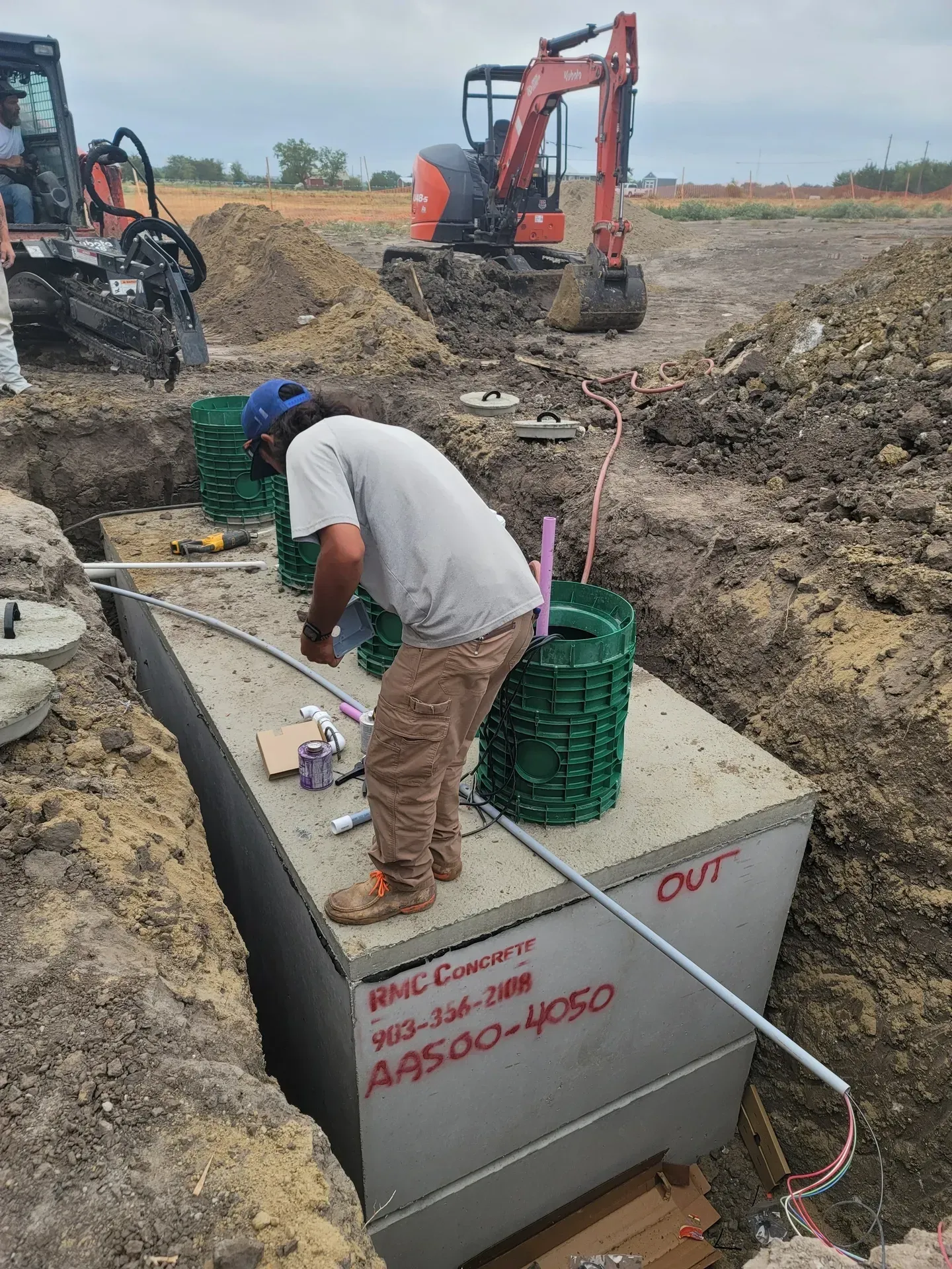 Worker installing conduit on a concrete septic tank in a construction site, with backhoe in background.