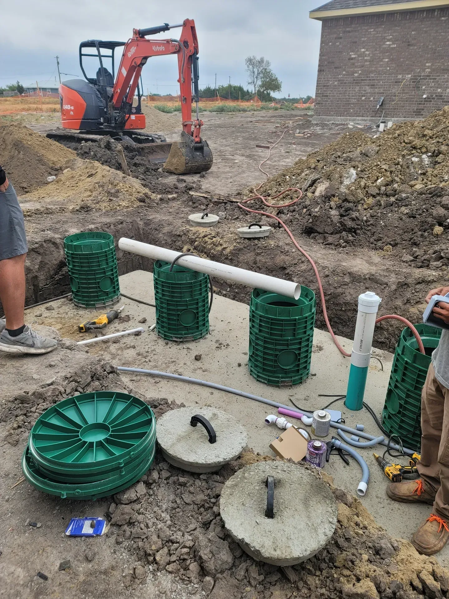 Construction site: installing septic tank components. Green cylinder cages, concrete lids, white PVC pipes, small excavator in background.