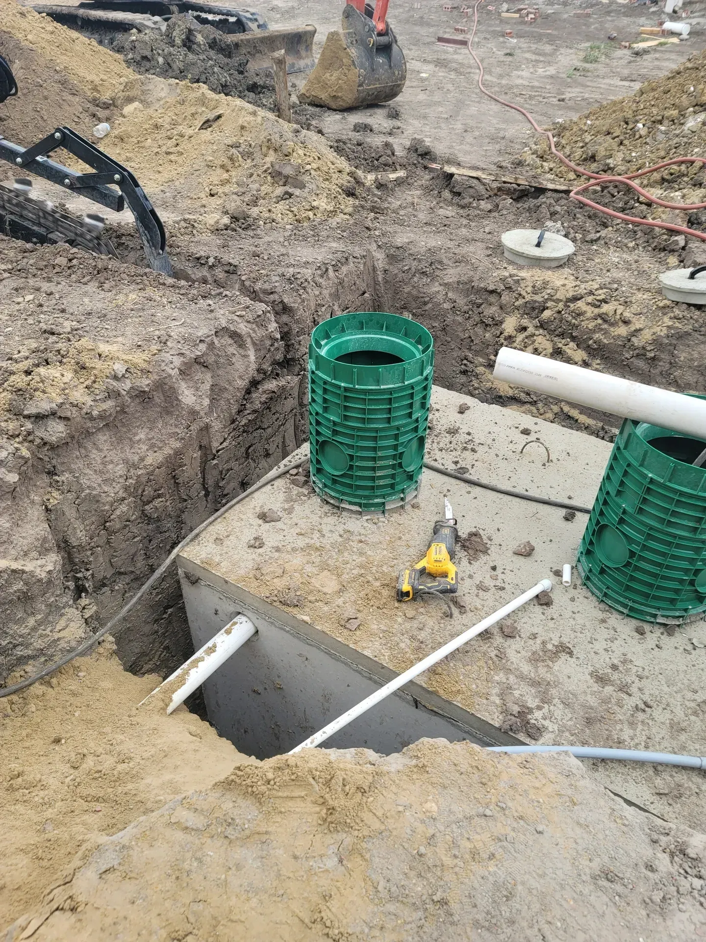 Septic tank installation in progress with exposed tank, piping, and green risers in a construction site.