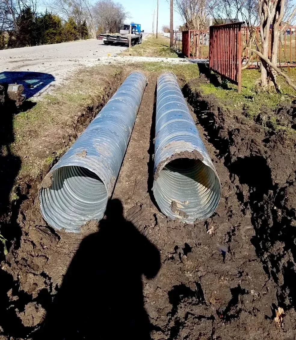 Two large corrugated metal pipes in trenches, alongside a road.