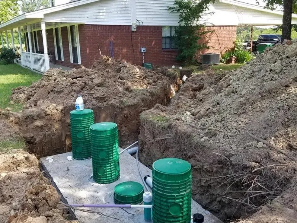 Septic system installation in progress: green tanks and pipes in excavated earth, near a house.