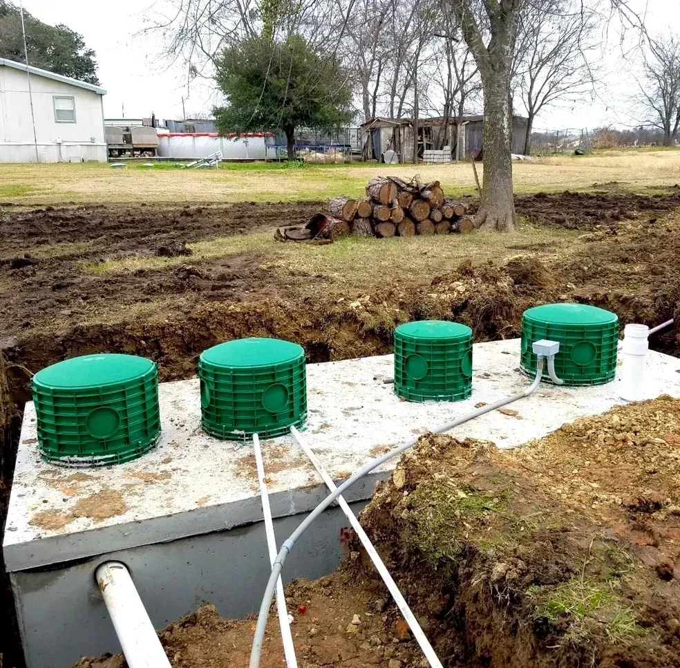 Septic system installation: green covers, concrete base, trenches in yard, rural setting.