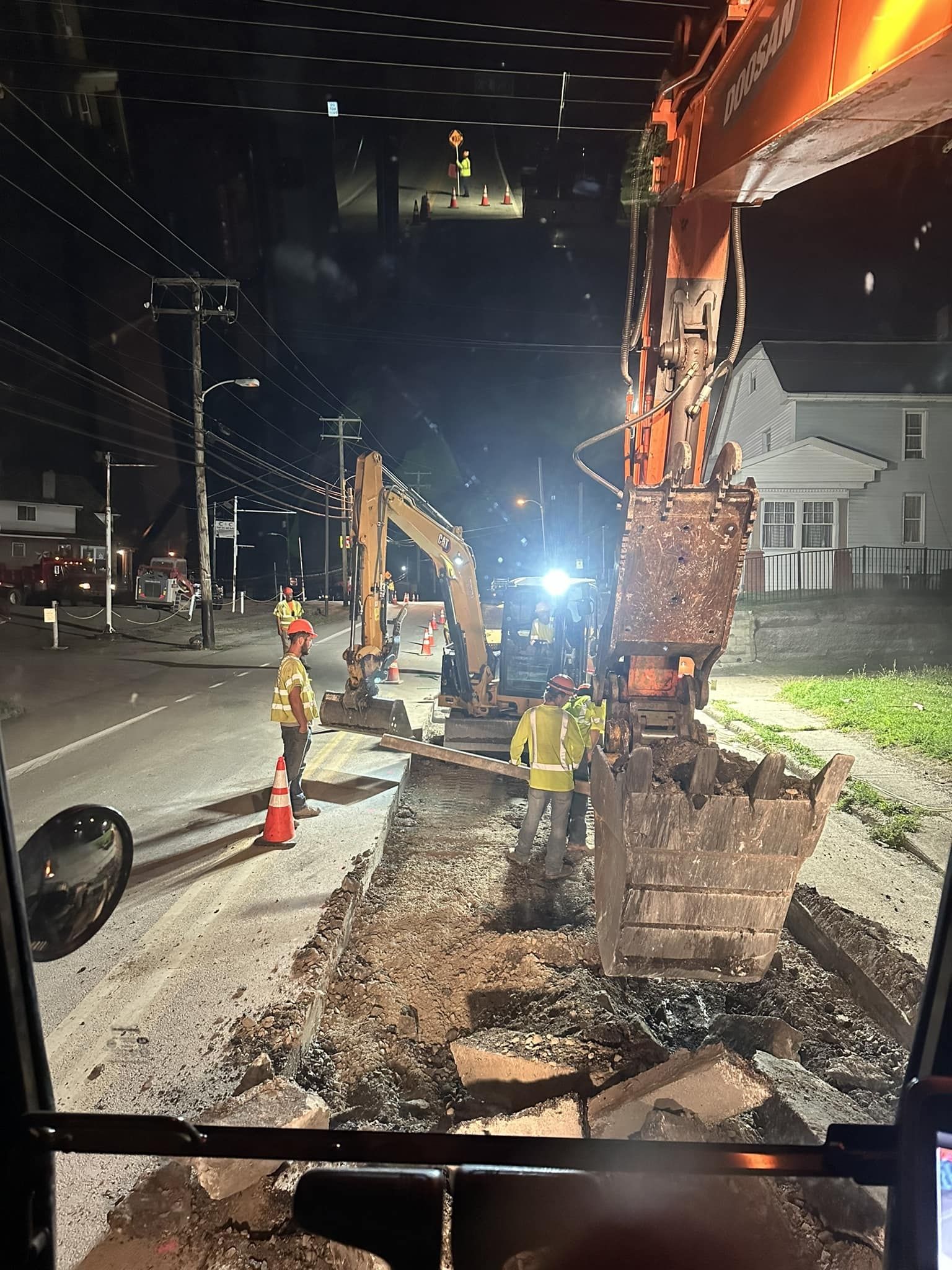 Nighttime road construction with two excavators and workers in high-visibility gear digging up a street.