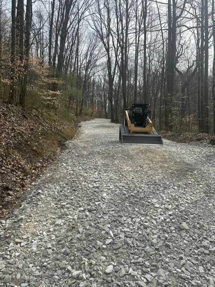 A yellow skid steer loader sits on a freshly graveled road winding through a forest with bare trees.