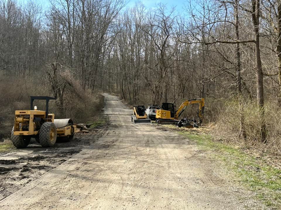 Construction equipment, including a roller and an excavator, parked on a dirt road in a wooded area.