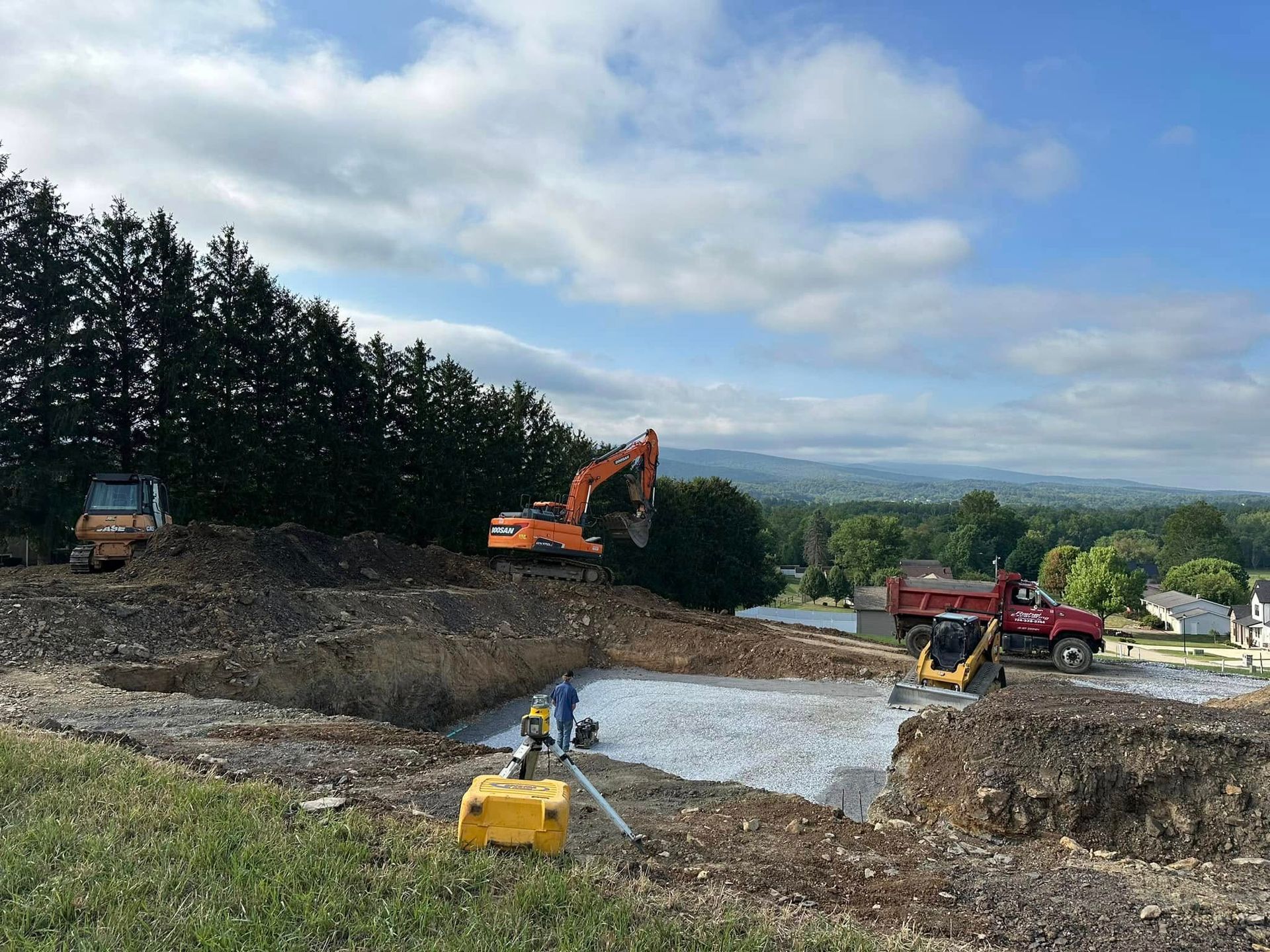 Construction equipment, including an excavator and a dump truck, works at a site with cleared dirt and a gravel base.