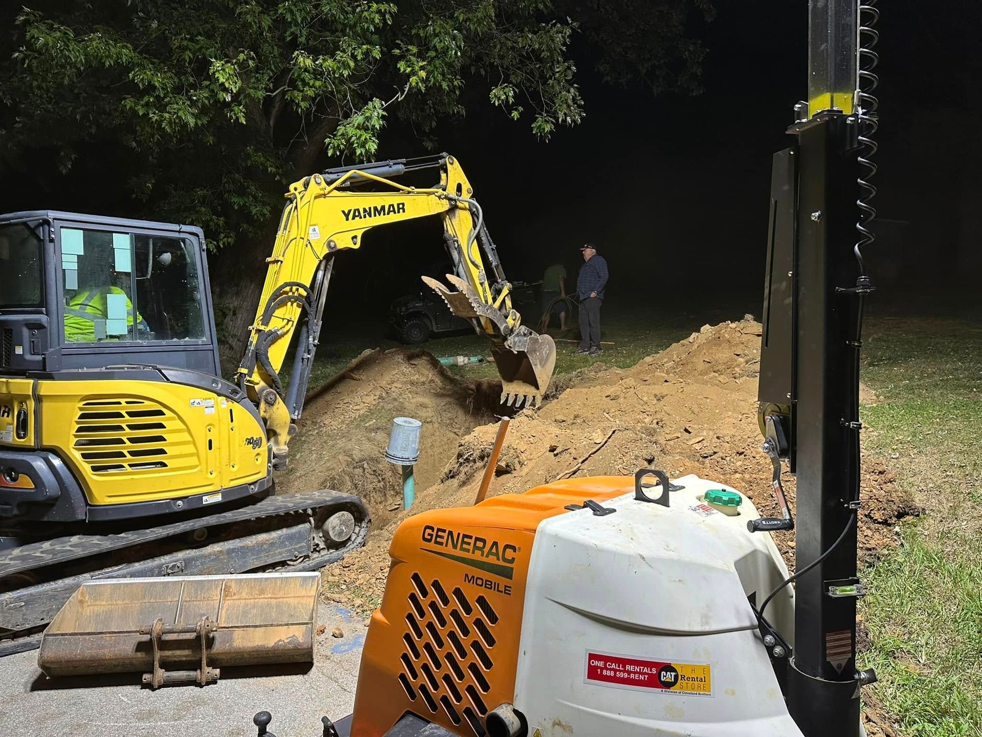 A yellow excavator and a Generac light tower at a construction site at night, with a mound of dirt and workers.