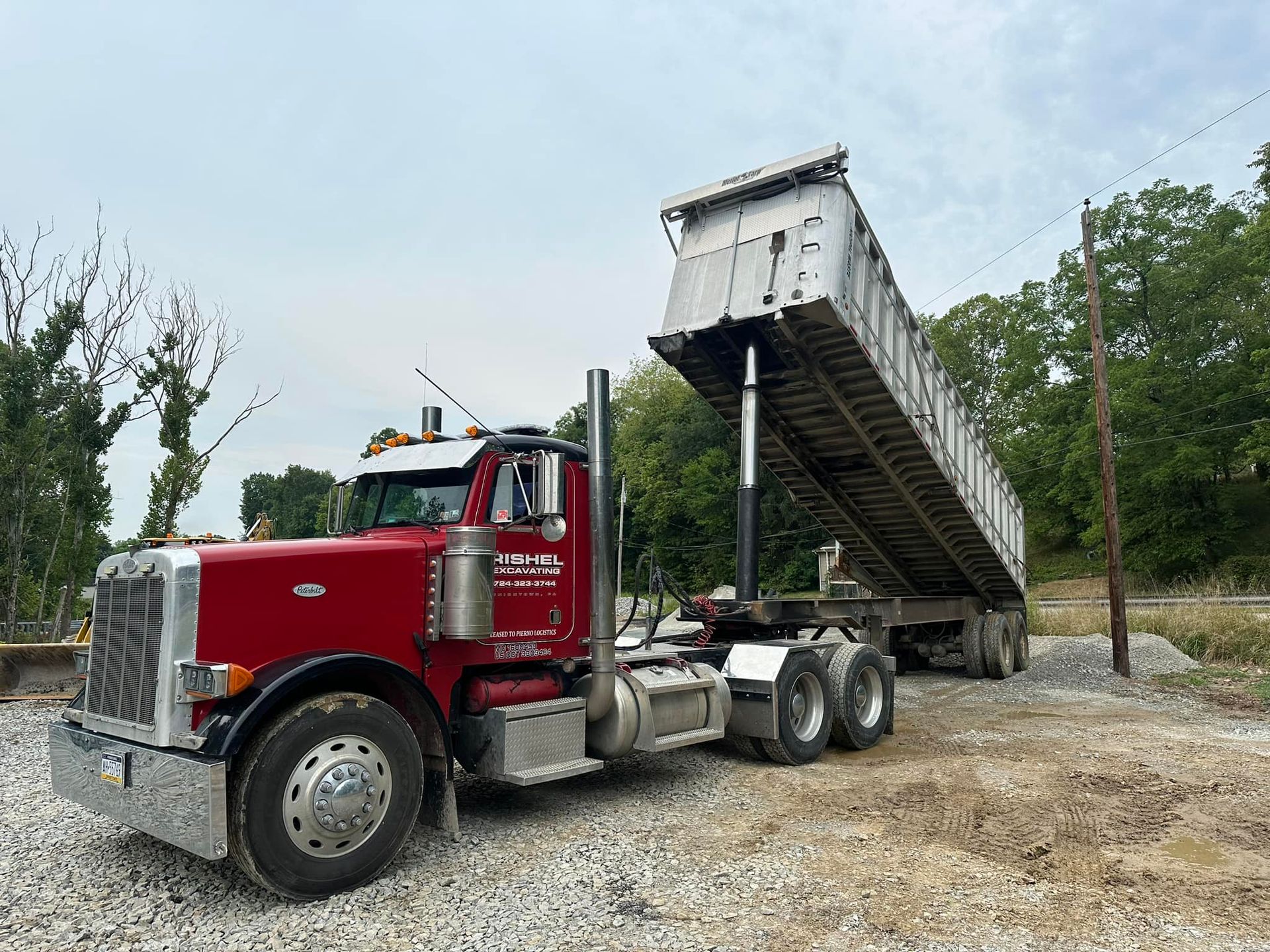 A red semi-truck with a silver dump trailer raised in the air, parked on a gravel lot near a tree line.